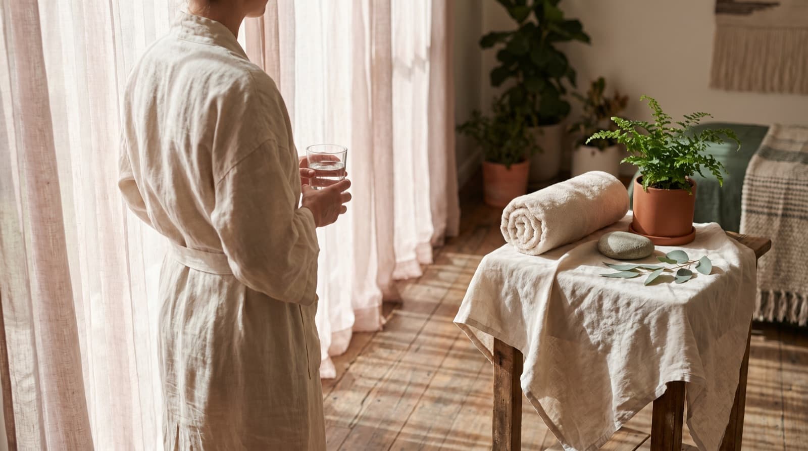 Fern & Soul massage aftercare in Innaloo — a client in a cream linen robe holding a glass of water by a sunlit window, beside a rolled towel and a potted fern.