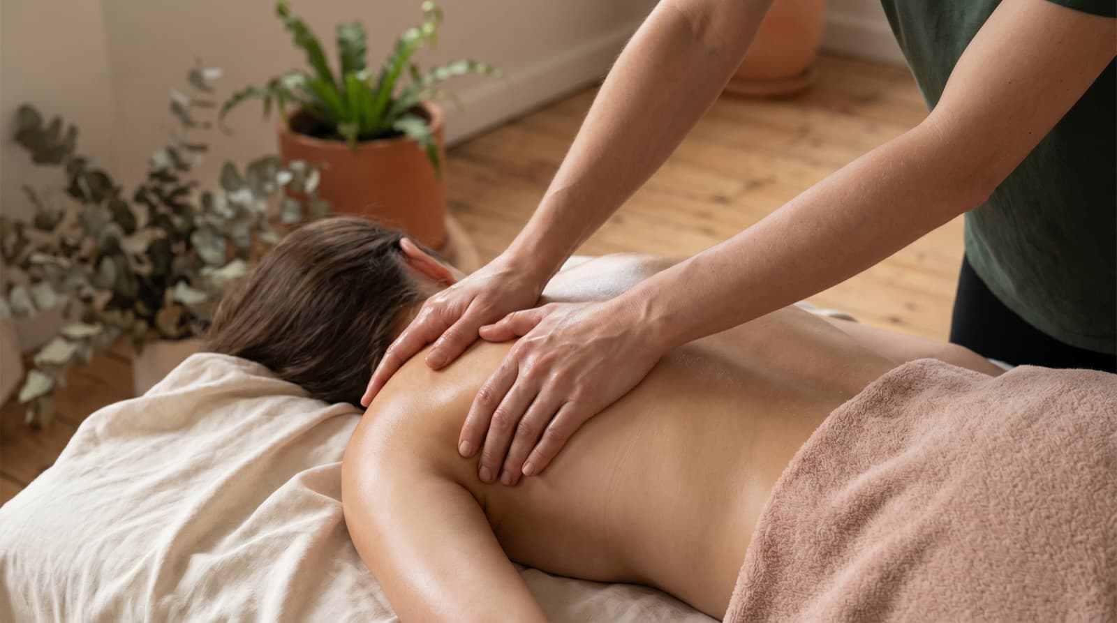 Remedial massage therapist using both hands to work on a relaxed client's upper back and shoulder on a linen-draped table.