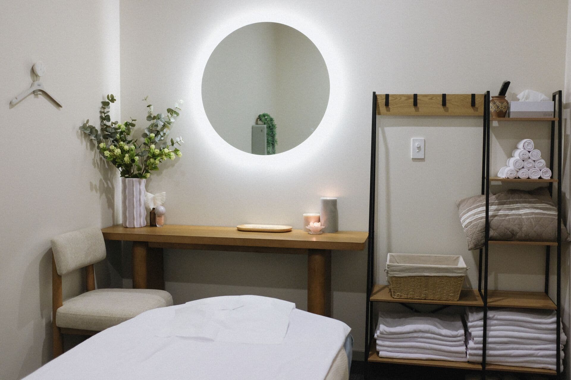 Backlit round mirror, eucalyptus on an oak console and shelves of folded linen beside a massage table at Fern & Soul in Innaloo.