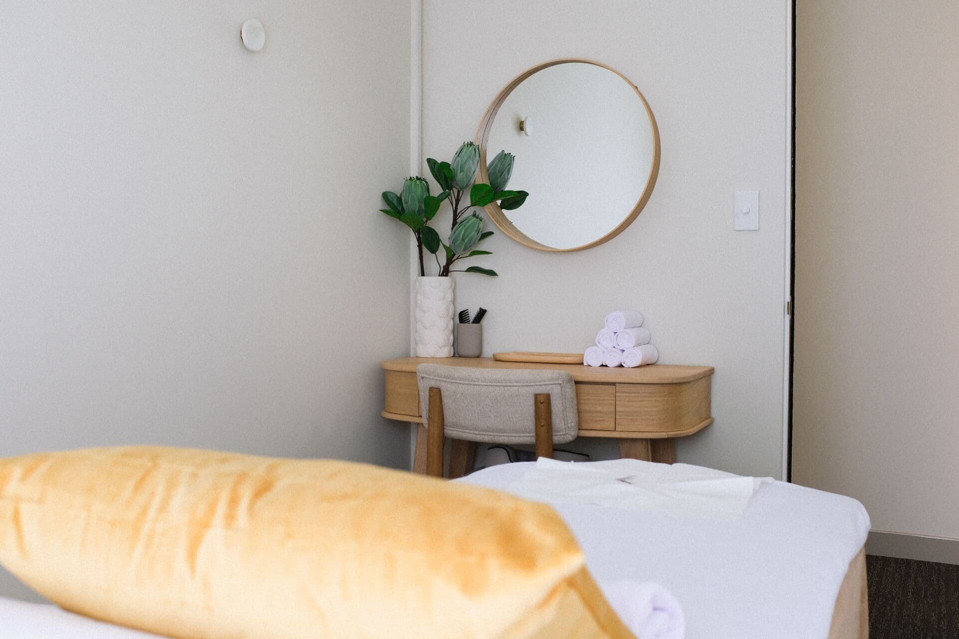 Velvet cushion on a made-up massage table in front of an oak vanity with a round mirror and rolled towels at Fern & Soul in Innaloo.
