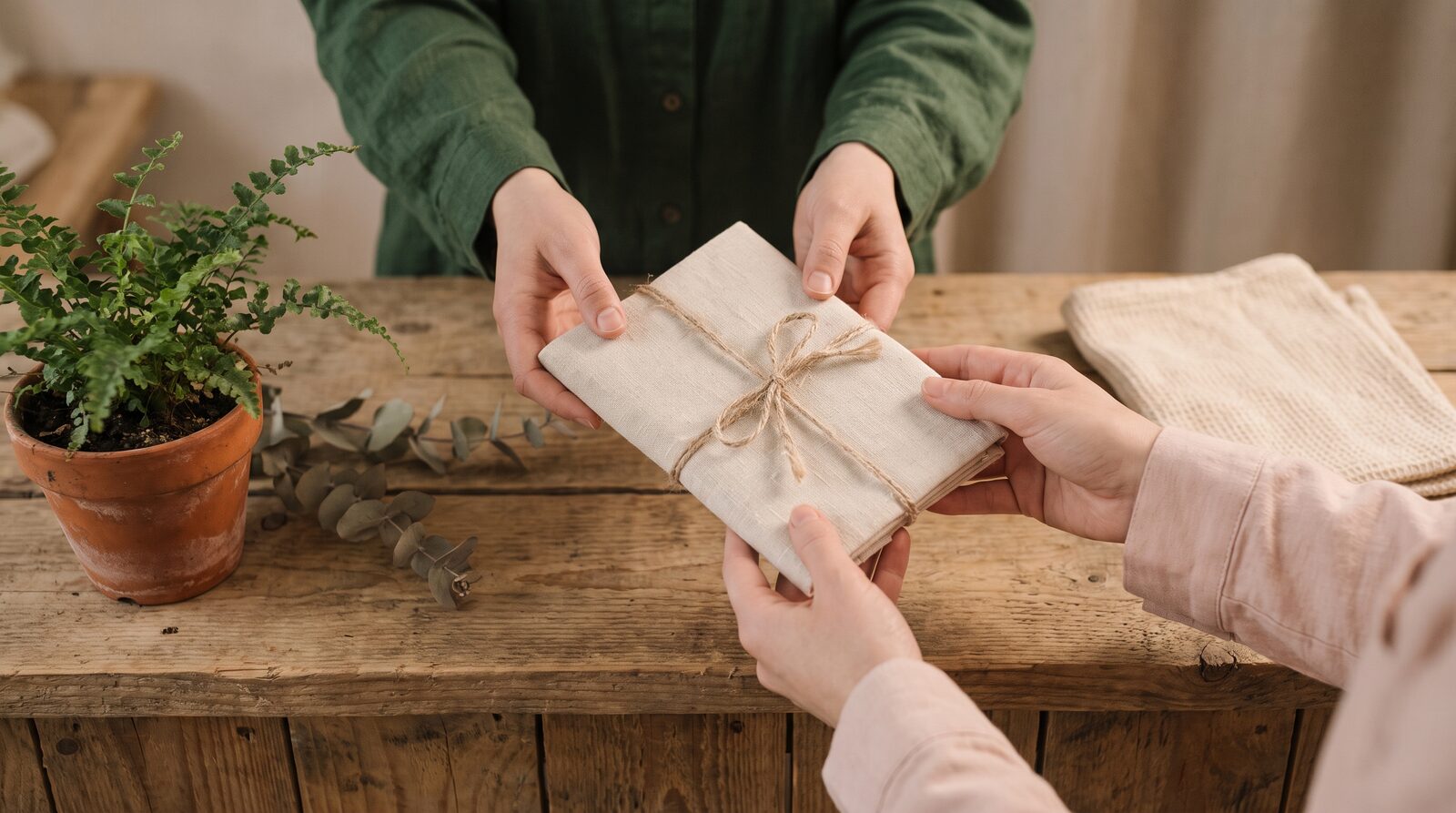 Fern & Soul massage gift voucher in Innaloo, hands passing a linen wrapped parcel tied with twine across a raw timber counter in warm light.